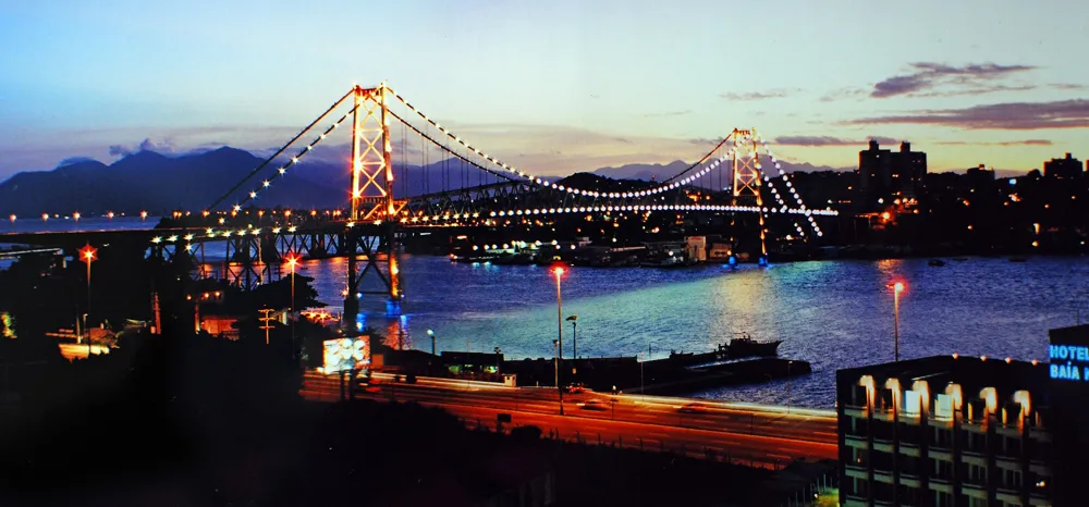 Silhueta da Ponte Hercílio Luz ao entardecer, céu alaranjado refletido na água